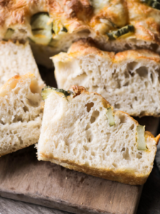 Sourdough pickle focaccia is shown on a wooden cutting board, cut into slices. You see the airy bubbles throughout the sourdough, the pickles, and fresh dill, and the crispy golden and cheesy crust.