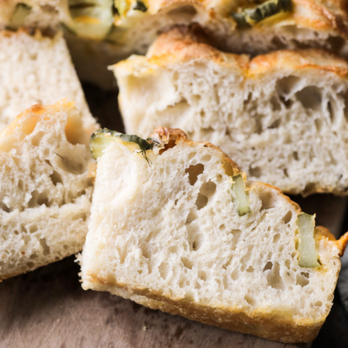Sourdough pickle focaccia is shown on a wooden cutting board, cut into slices. You see the airy bubbles throughout the sourdough, the pickles, and fresh dill, and the crispy golden and cheesy crust.