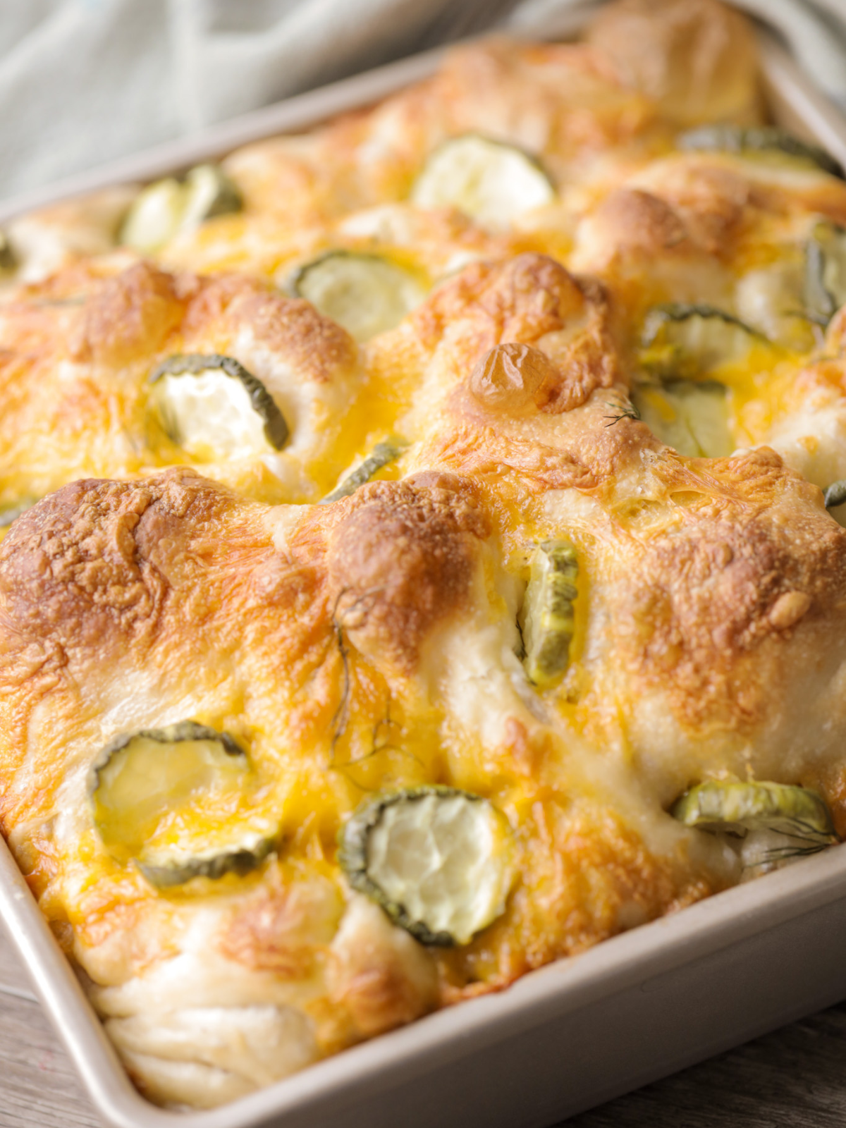 A closeup of the freshly baked sourdough pickle focaccia is shown. It has a golden, bubbly crust, melty shredded cheddar cheese, pickle chips, and fresh dill on top.