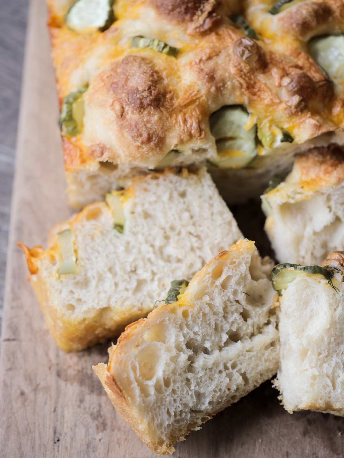 Several slices of sourdough dill pickle focaccia sit on a wooden cutting board, with the rest of the focaccia in the background.