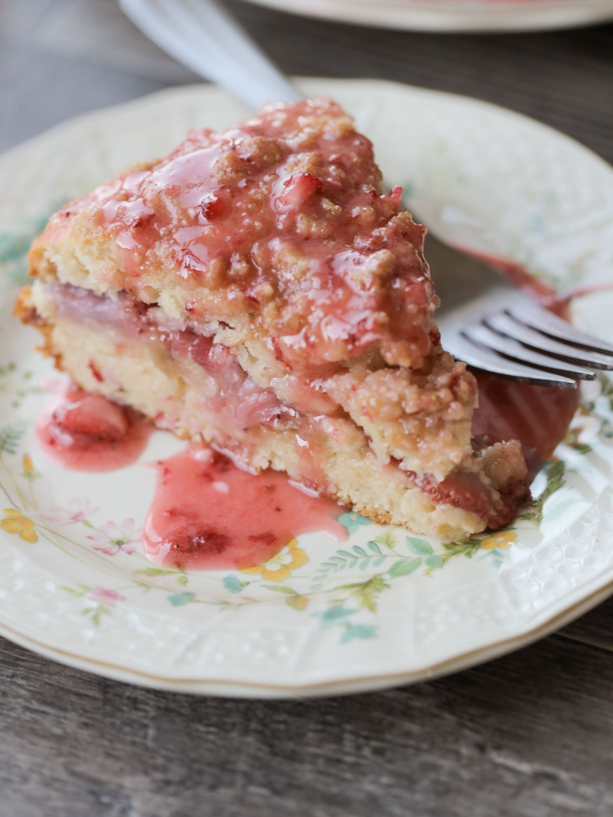 A slice of strawberry sourdough coffee cake sits on a floral plate with a fork. It is drizzled with fresh strawberry icing.