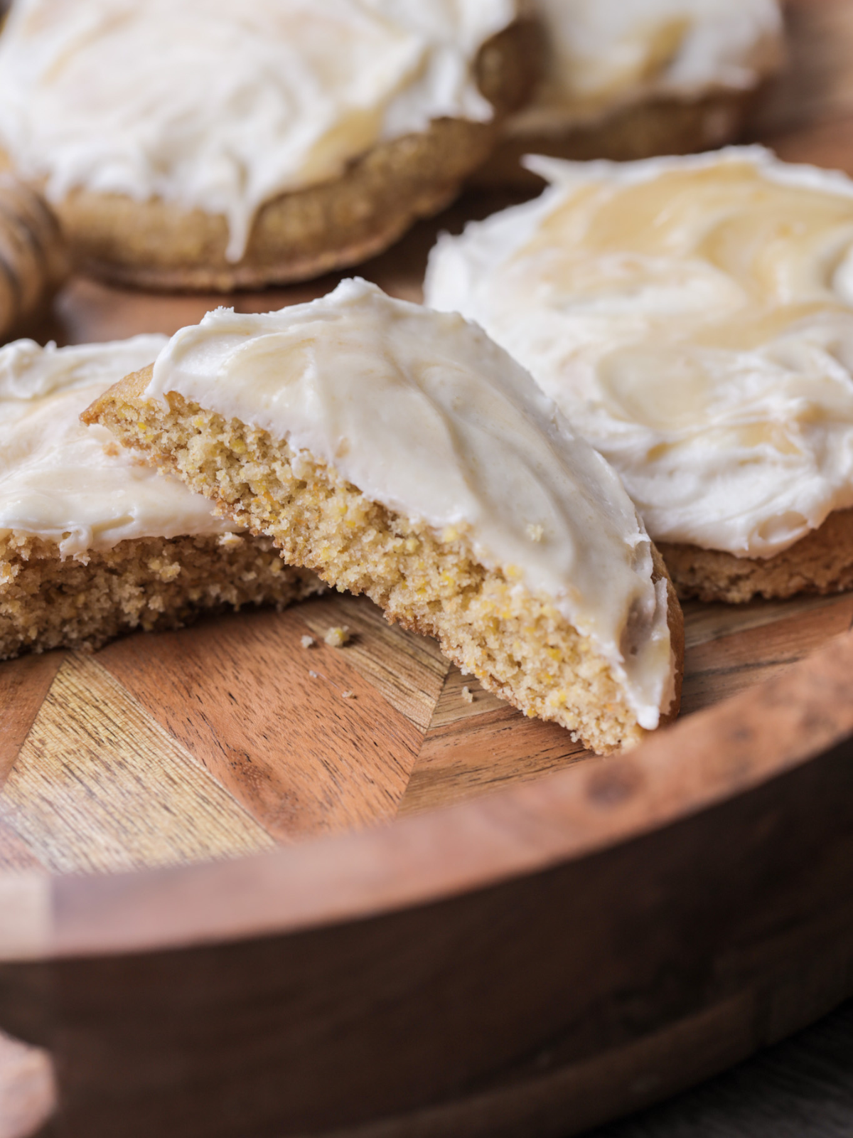 The frosted sourdough cornbread brown butter cookies sit on a wooden platter. There is one cookie cut in half, so you can see the moist, soft crumb of the cookie.