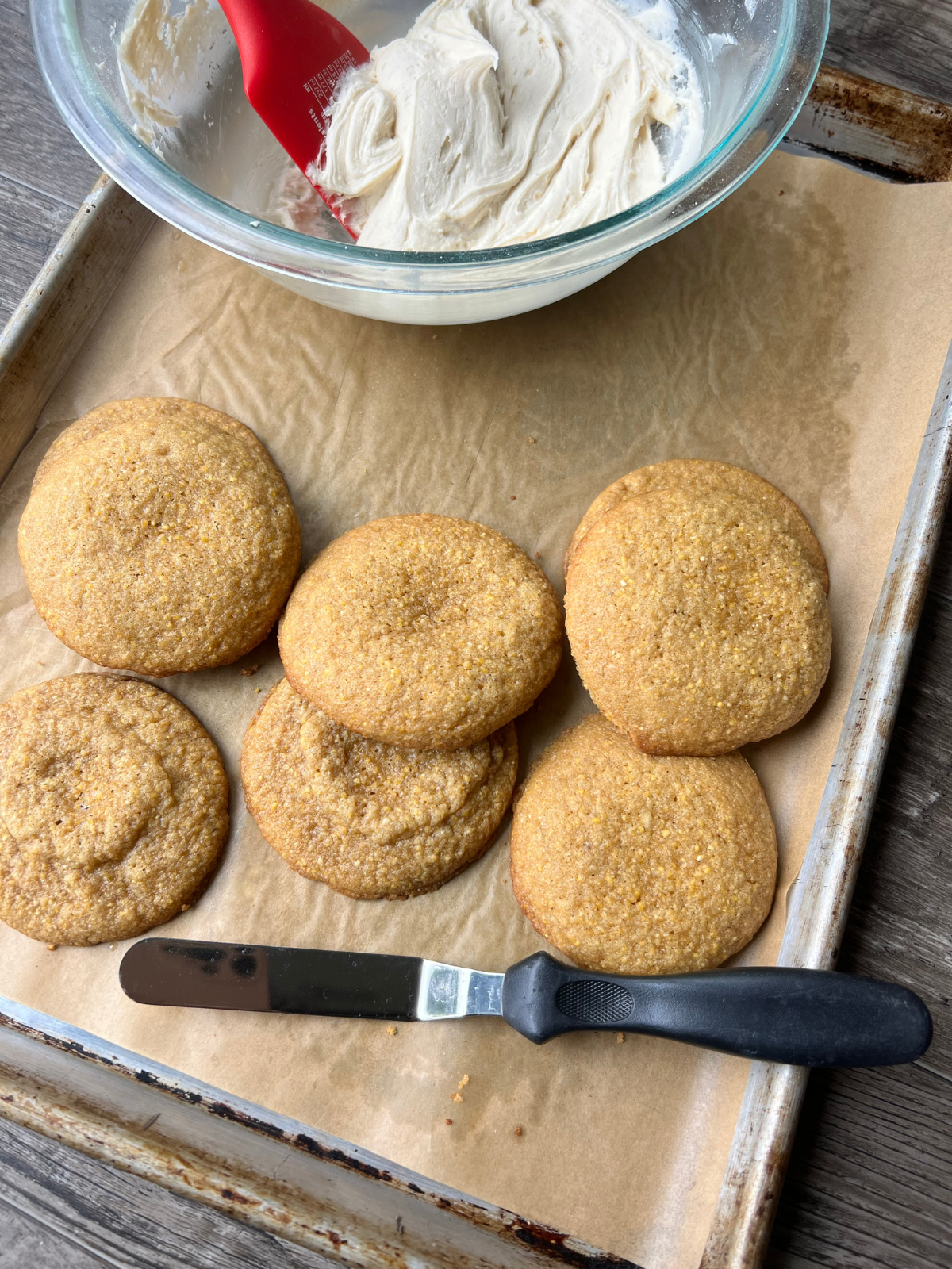 The freshly baked cornbread sourdough cookies sit on a baking sheet with the bowl of honey buttercream frosting behind them.
