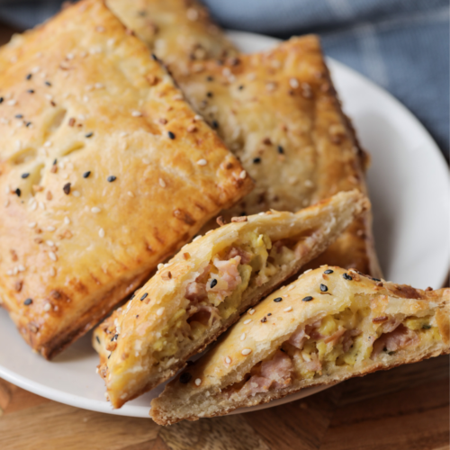 Three sourdough discard hand pies with ham, cheese, and egg filling sit on a plate. Another hand pie is cut in half so you can see the melty cheese and ham.