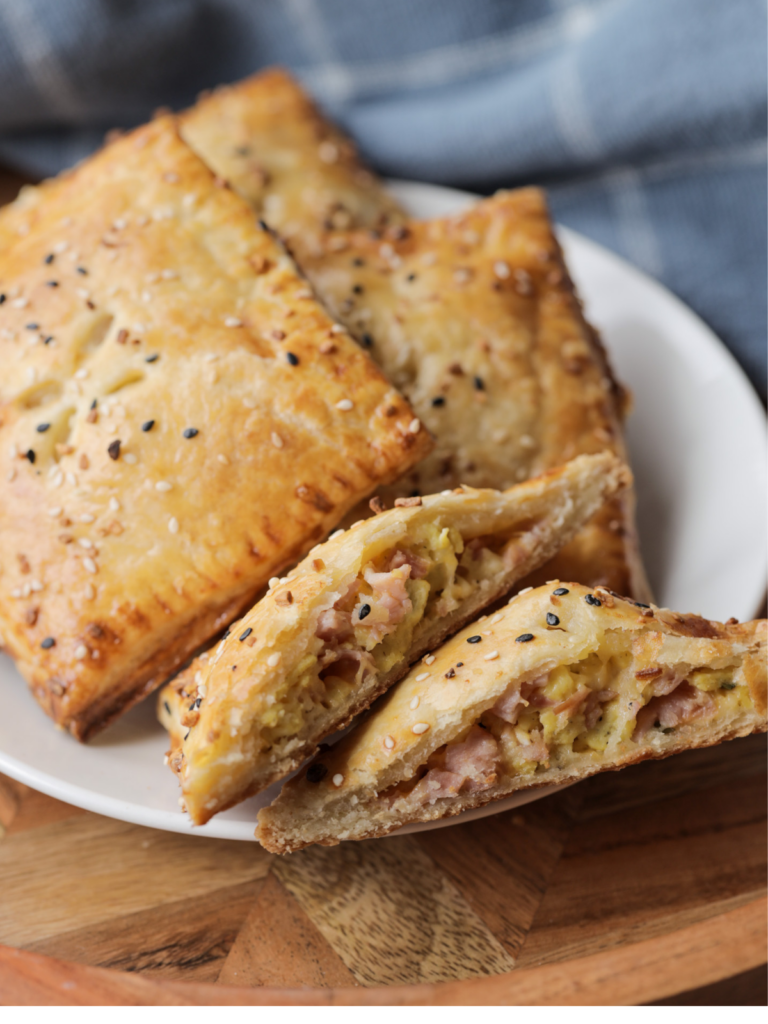 Three sourdough discard hand pies with ham, cheese, and egg filling sit on a plate. Another hand pie is cut in half so you can see the melty cheese and ham.