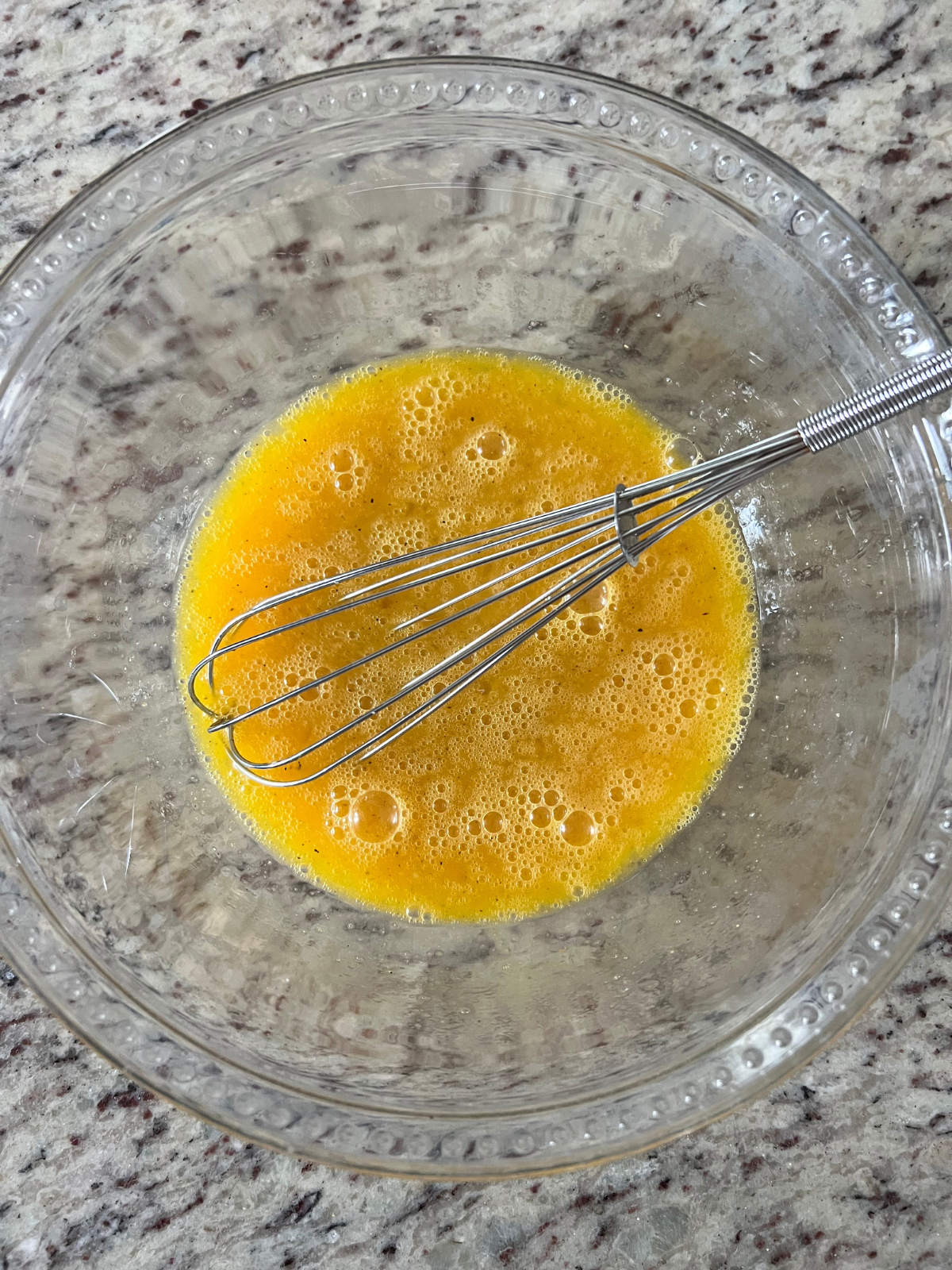 Whisked eggs, garlic salt, and pepper are shown in a glass mixing bowl with a whisk in it, preparing to be cooked on the skillet.