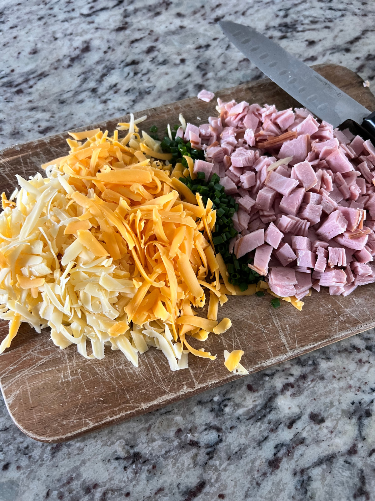 The filling ingredients for the sourdough discard hand pies (shredded smoked gouda, shredded sharp cheddar cheese, chopped chives, diced ham) are shown on a wooden cutting board.