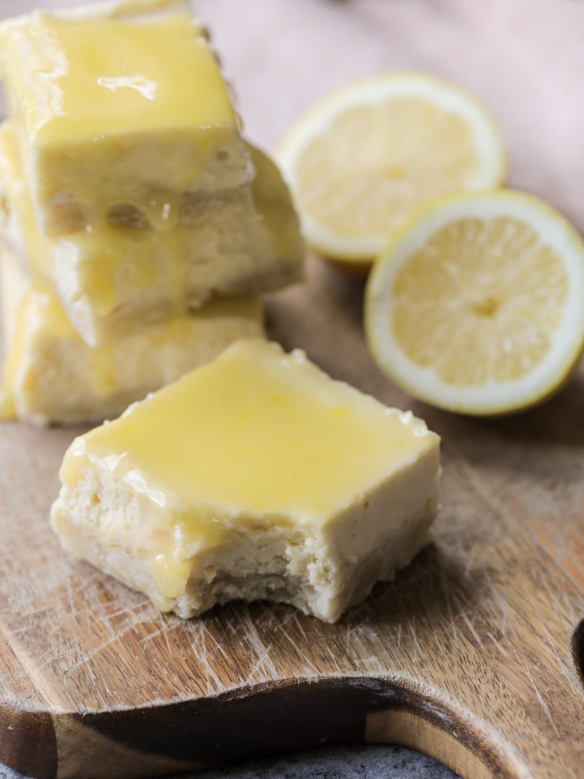 A lemon curd cheesecake bar is shown with a bite taken out of it. Three more cheesecake bars are stacked behind it with two fresh lemon halves in the background.