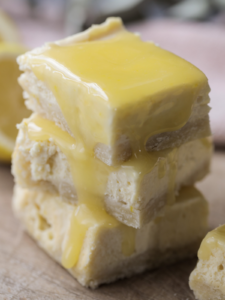 Three lemon curd cheesecake bars with sourdough shortbread crust sit stacked on a wooden cutting board. Lemon curd drips down the sides of the bars.
