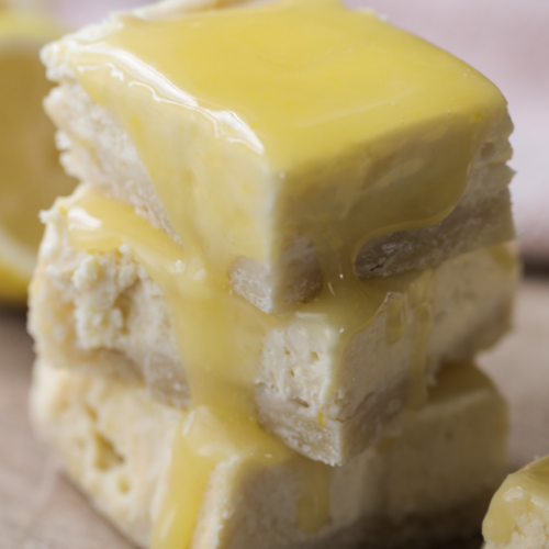 Three lemon curd cheesecake bars with sourdough shortbread crust sit stacked on a wooden cutting board. Lemon curd drips down the sides of the bars.