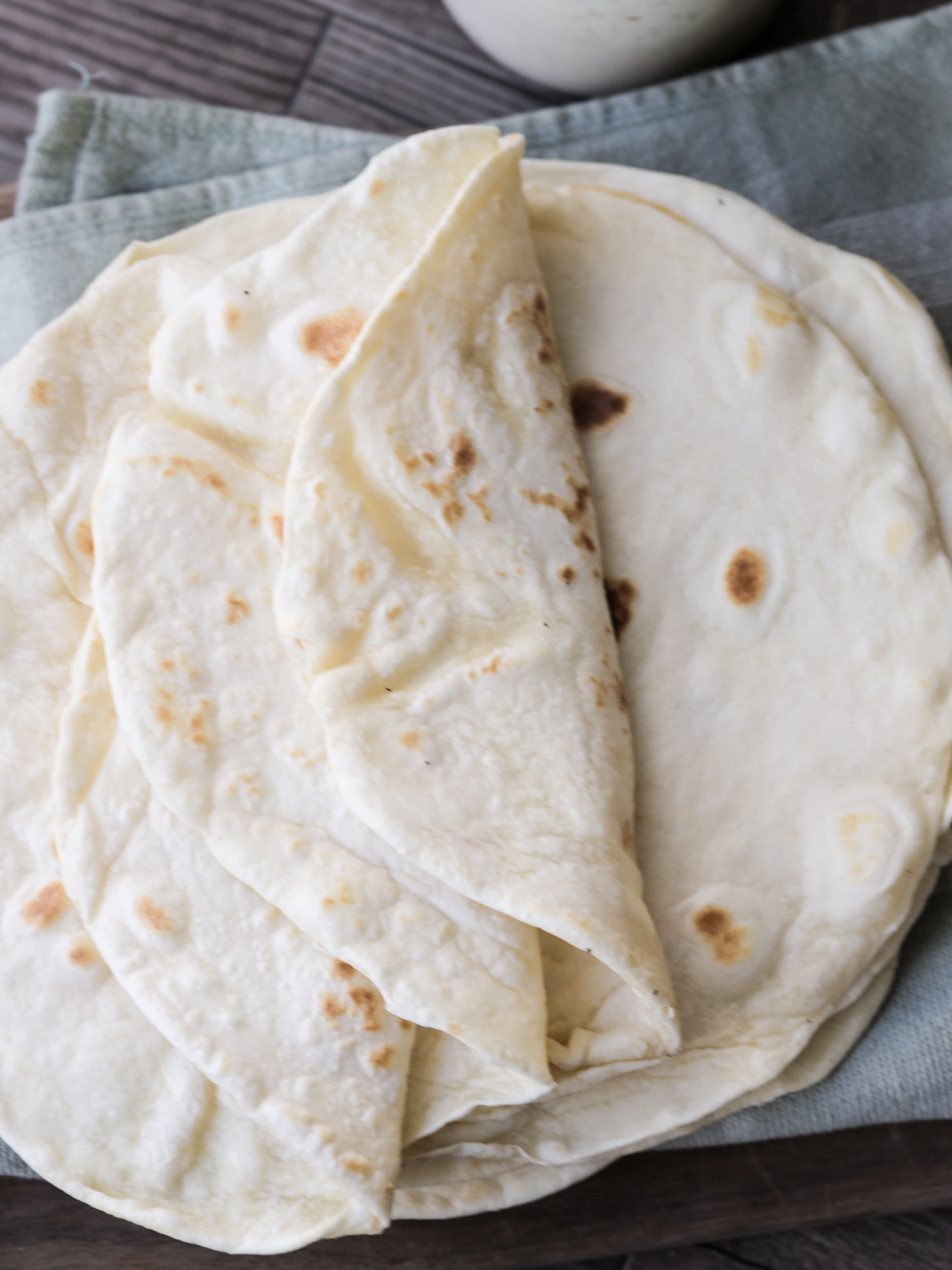 Several sourdough tortillas sit on a kitchen towel. A few of the top tortillas are shown folded over to show the softness.