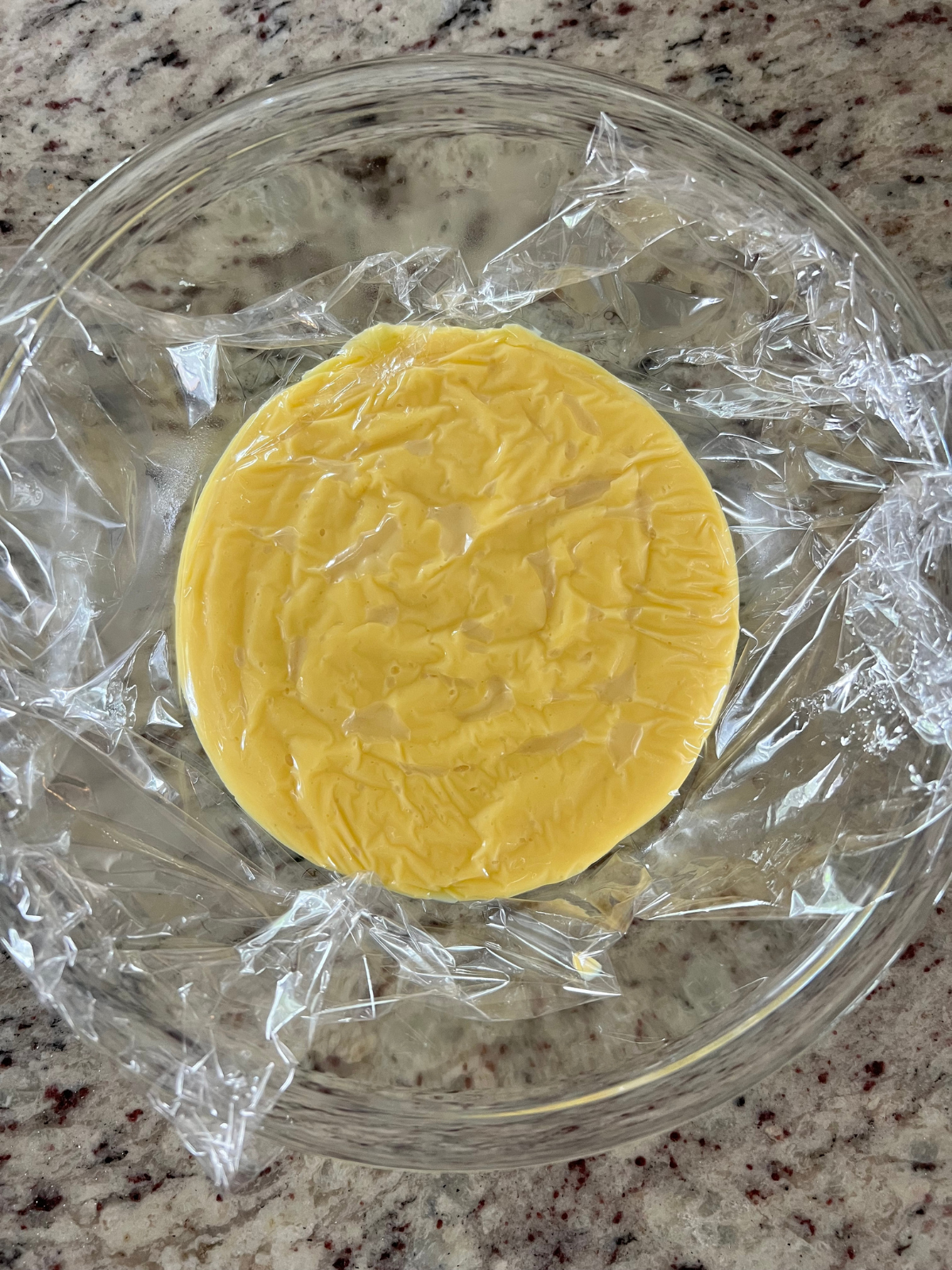 The vanilla custard sits in a glass mixing bowl with plastic wrap laying over it.