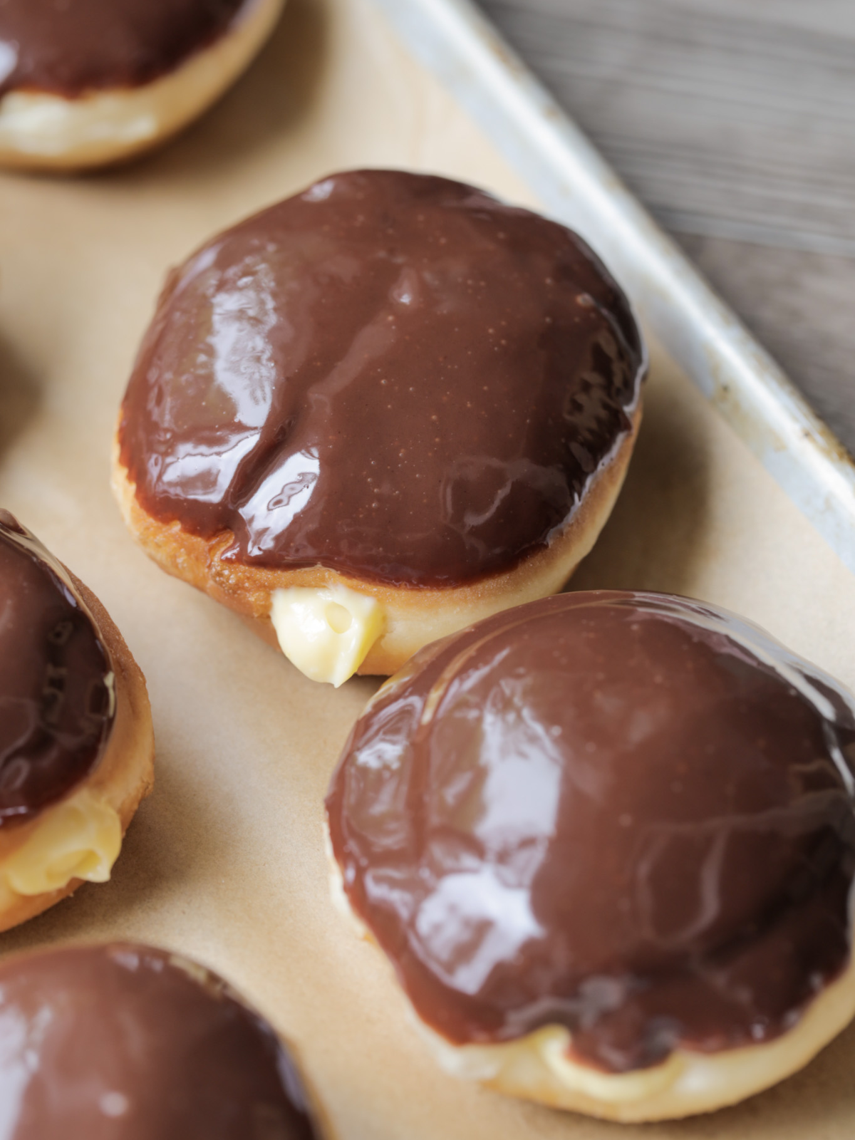 A closeup of one sourdough Boston cream donut sits on a baking sheet with several others surrounding it.