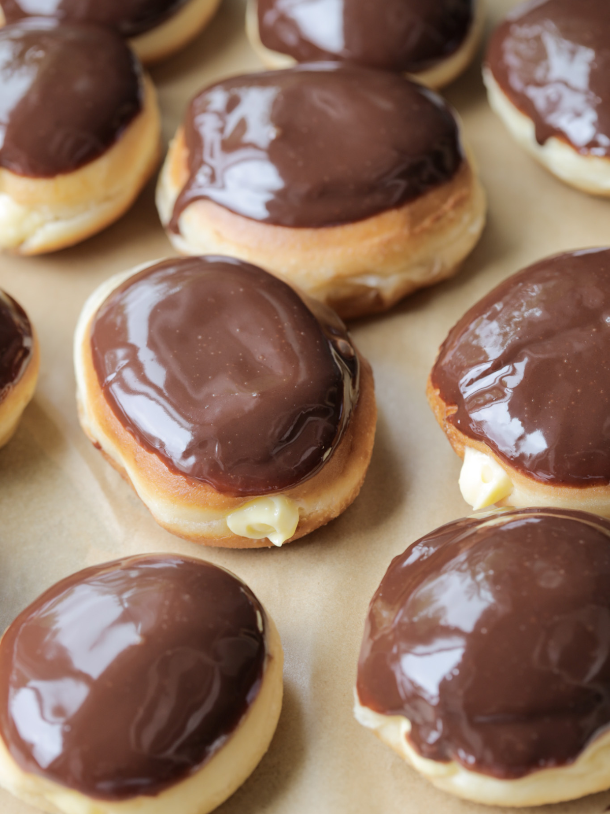 Twelve sourdough brioche Boston cream donuts with vanilla custard sit on a baking sheet. They are topped with a homemade and simple chocolate ganache.
