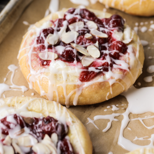 A closeup of a sourdough cream cheese bun with icing drizzled over it and several other sweet buns lay next to it.