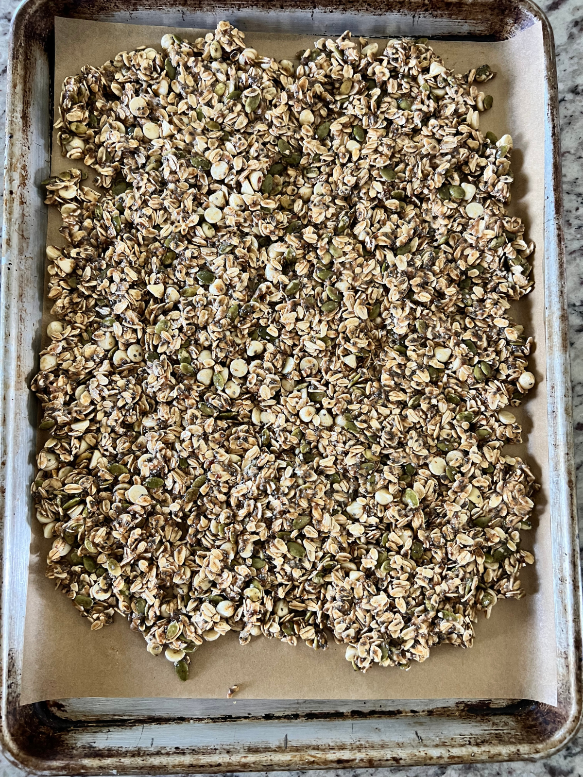 The sourdough white chocolate granola is shown spread out over a parchment paper-lined baking sheet preparing to be baked.