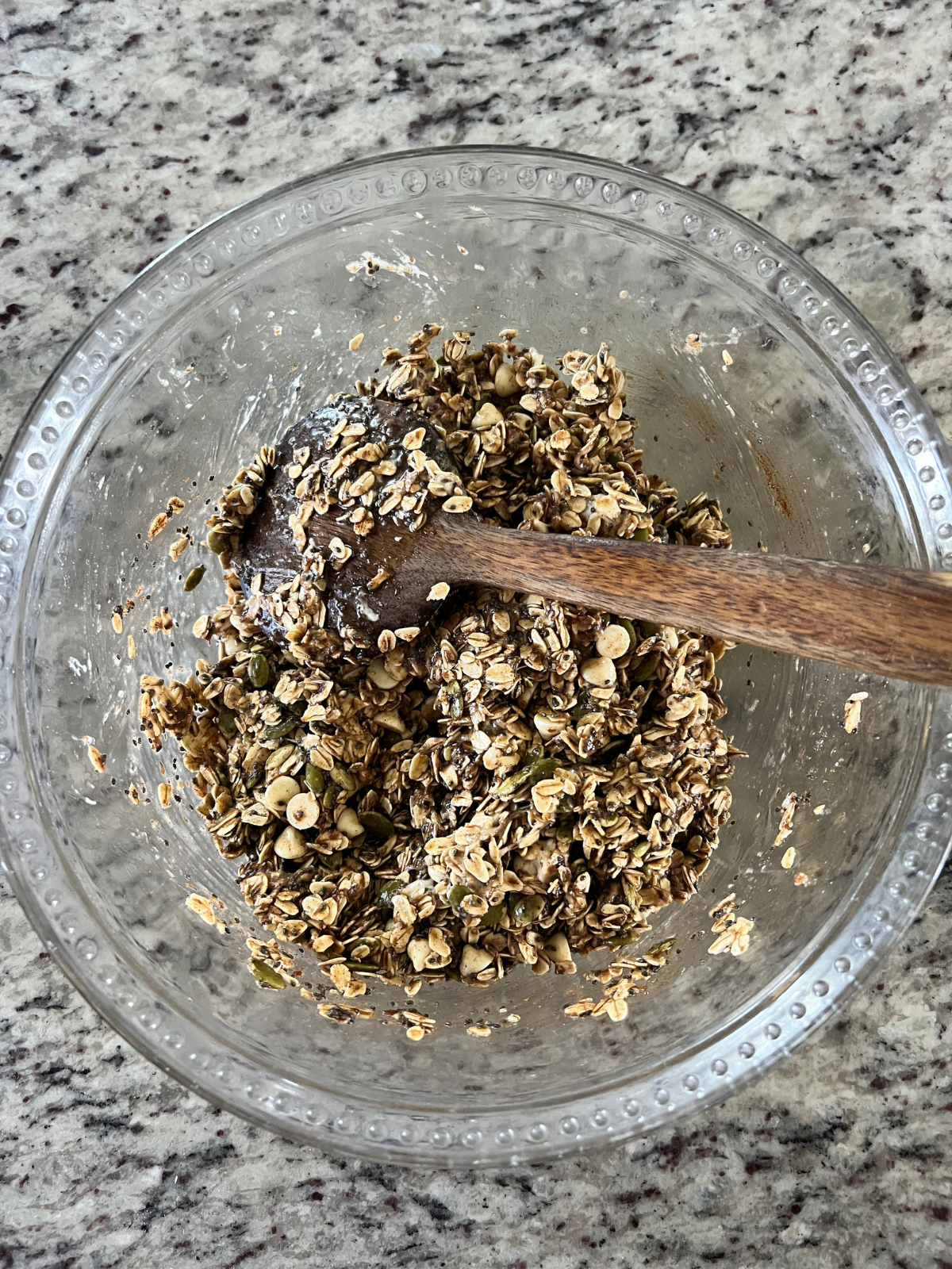 The ingredients for the sourdough granola is shown mixed together in a glass mixing bowl with a wooden spoon resting in it. 