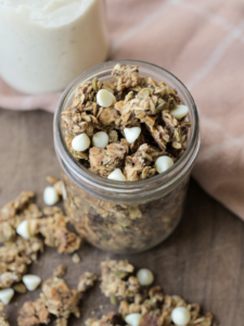 White chocolate sourdough discard granola sits in a glass jar with granola laying on the wooden platter below the jar.