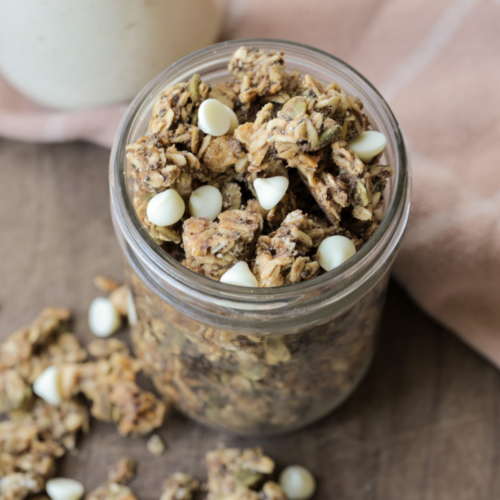 White chocolate sourdough discard granola sits in a glass jar with granola laying on the wooden platter below the jar.