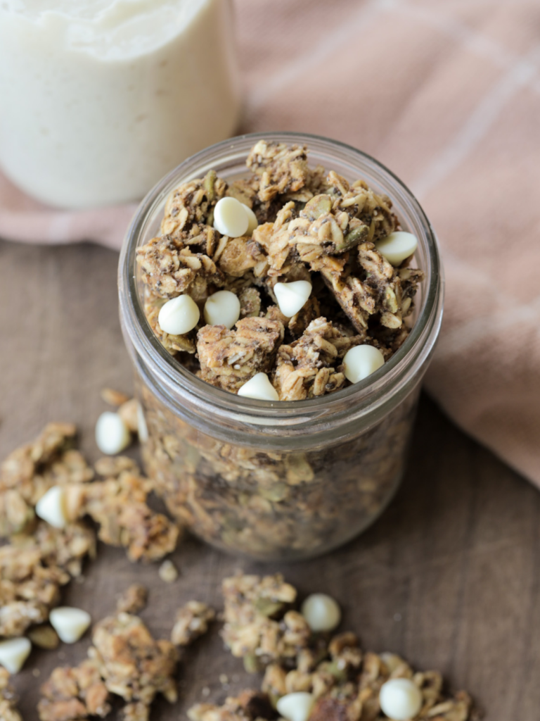 White chocolate sourdough discard granola sits in a glass jar with granola laying on the wooden platter below the jar.