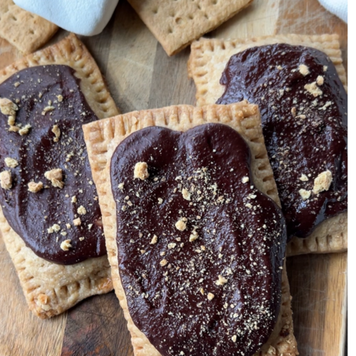 Three sourdough smores pop tarts sit on a wooden cutting board with marshmallows and graham crackers in the background.