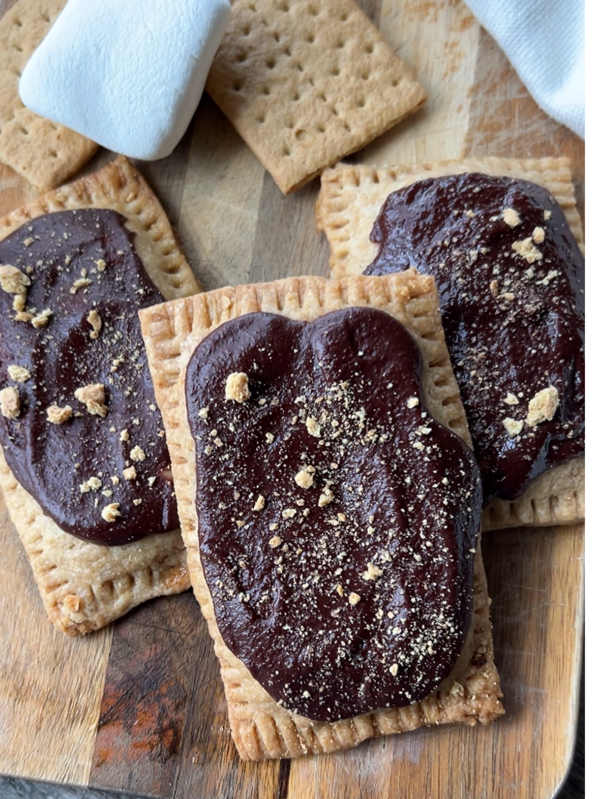 Three sourdough smores pop tarts sit on a wooden cutting board with marshmallows and graham crackers in the background.