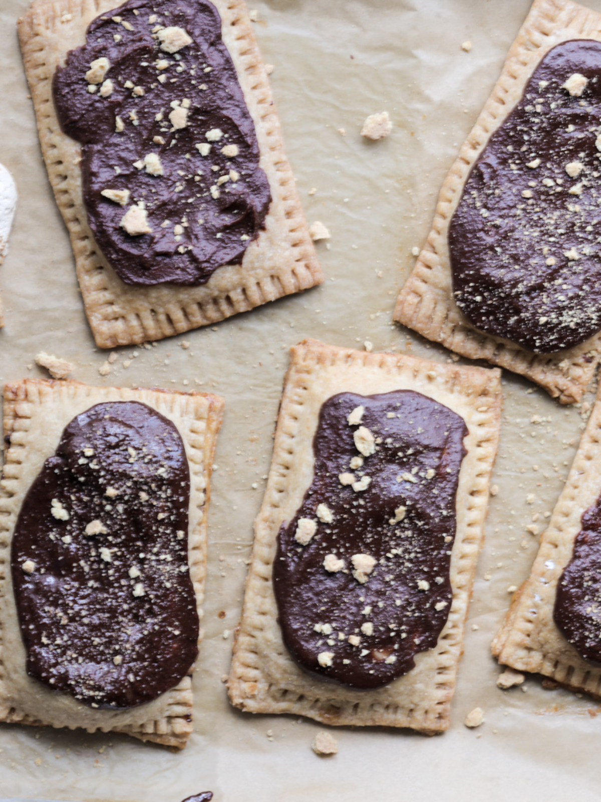 Several sourdough s'mores pop tarts rest on a baking sheet after being topped with chocolate icing and crushed graham crackers.