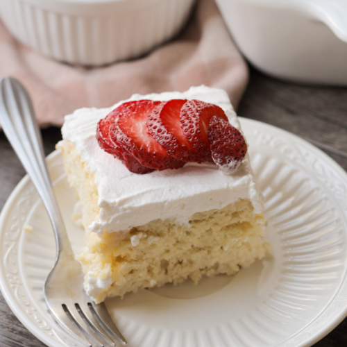 A slice of sourdough discard tres leches cake with whipped cream frosting and sliced strawberries rests on a plate with a fork on it. The full sourdough tres leches cake sits in the background along with a bowl of strawberries.
