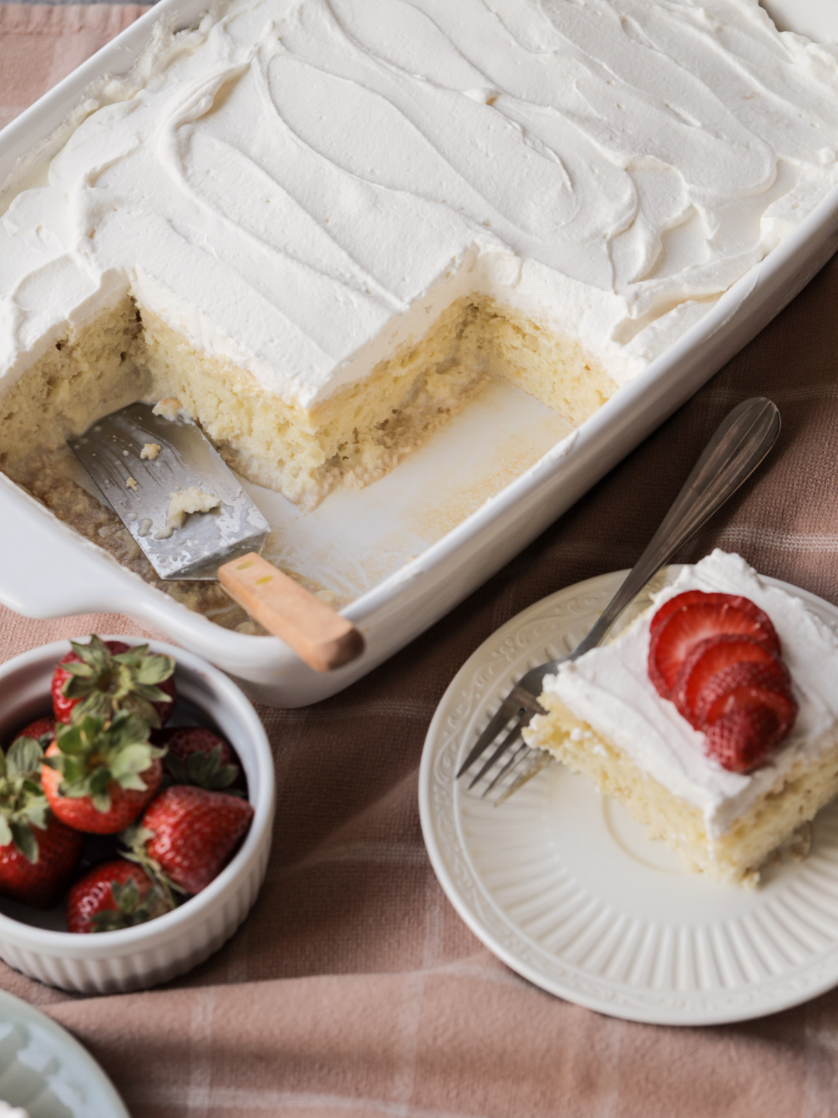 A full sourdough discard tres leches cake (with a few pieces taken out) and a slice of cake with sliced strawberries rest on the counter. A bowl of strawberries sit beside them.