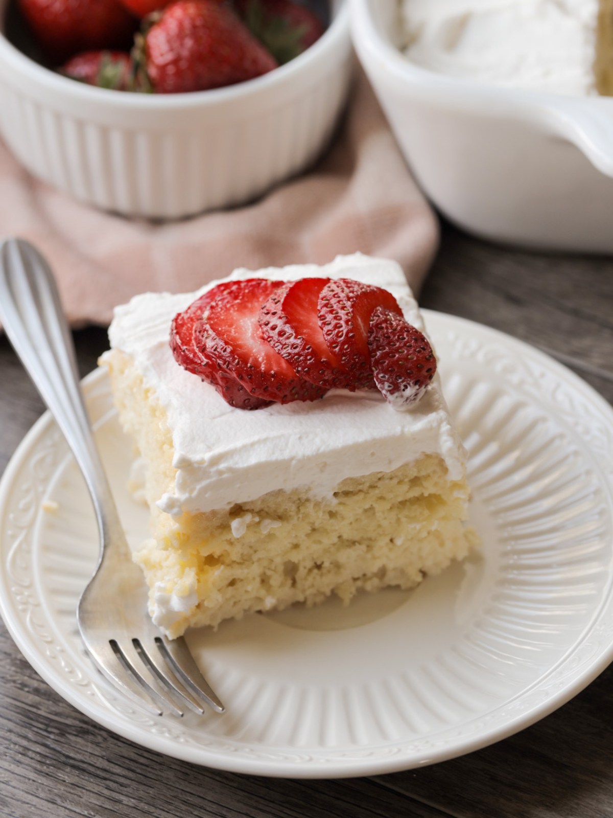 A slice of sourdough discard tres leches cake with whipped cream frosting and sliced strawberries rests on a plate with a fork on it. The full sourdough tres leches cake sits in the background along with a bowl of strawberries.