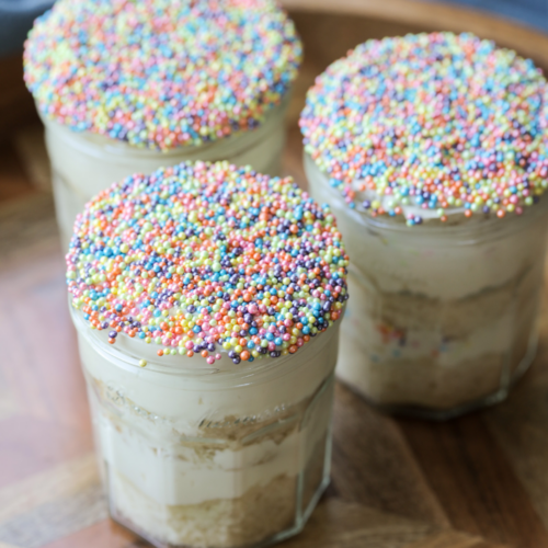 Three sourdough vanilla dot cakes are shown with vanilla cream cheese frosting in glass jelly jars. They are topped with a layer of dot sprinkles.