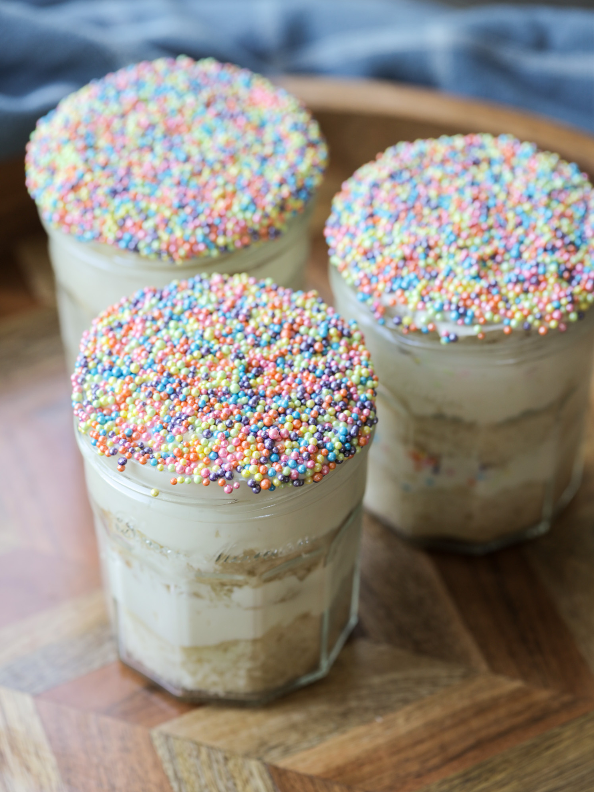 Three sourdough vanilla dot cakes are shown with vanilla cream cheese frosting in glass jelly jars. They are topped with a layer of dot sprinkles.