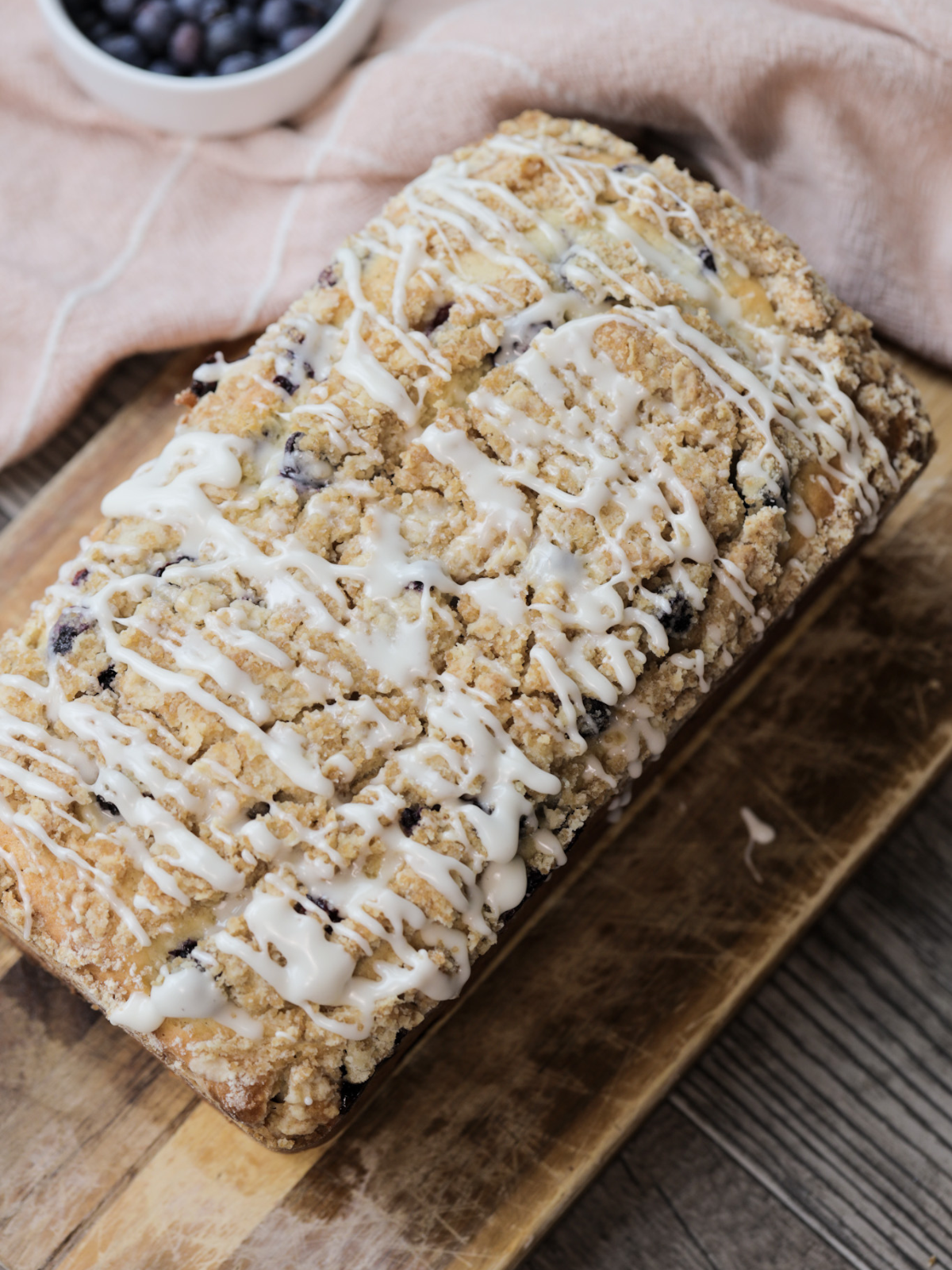 The sourdough discard blueberry loaf with icing drizzled over it sits on a wooden cutting board.