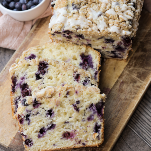 A loaf of sourdough blueberry crumble with glaze sits on a wooden cutting board. It has two slices cut out, showing the bursts of fresh blueberries throughout.