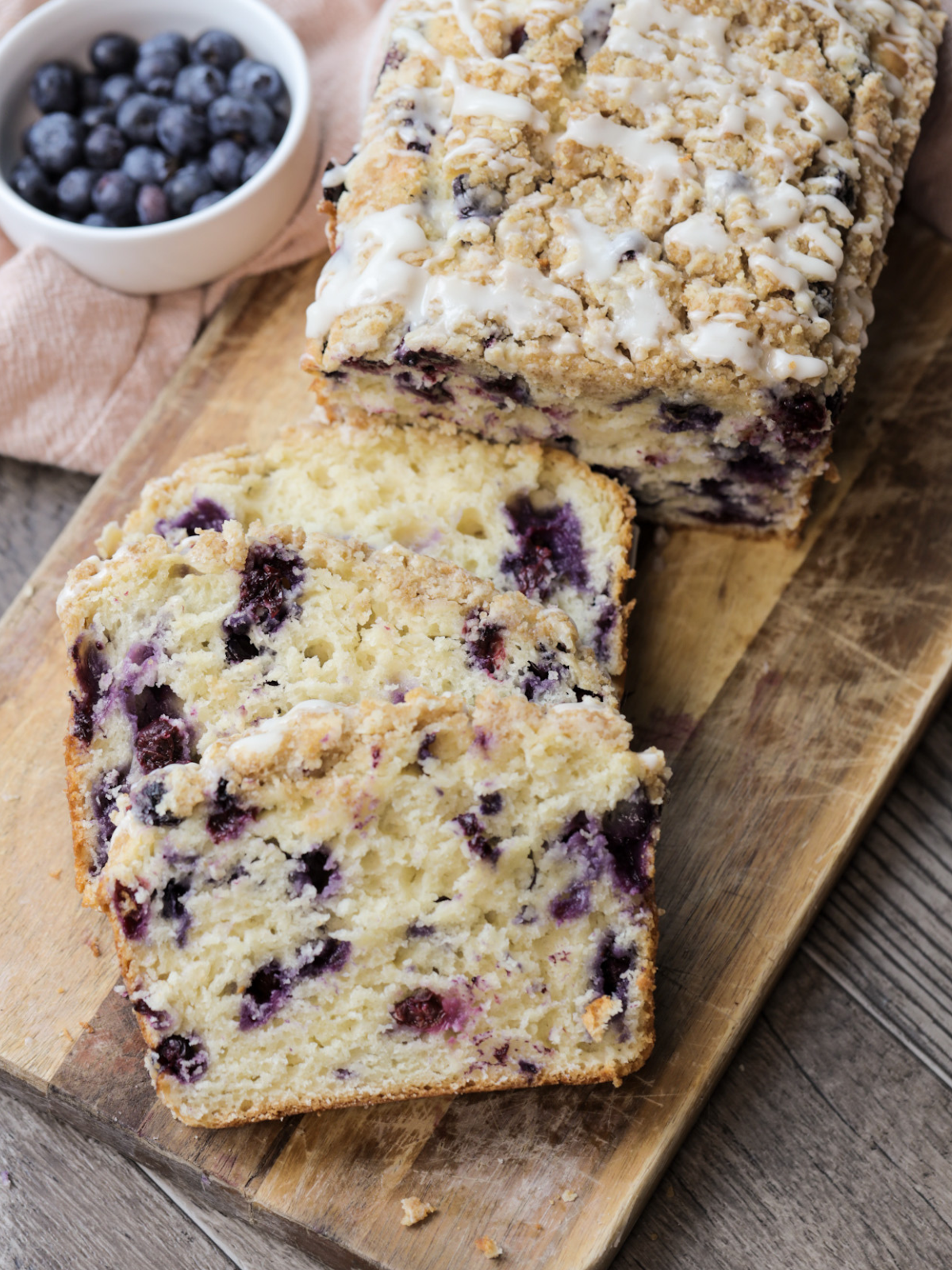 A loaf of sourdough blueberry crumble with glaze sits on a wooden cutting board. It has two slices cut out, showing the bursts of fresh blueberries throughout.