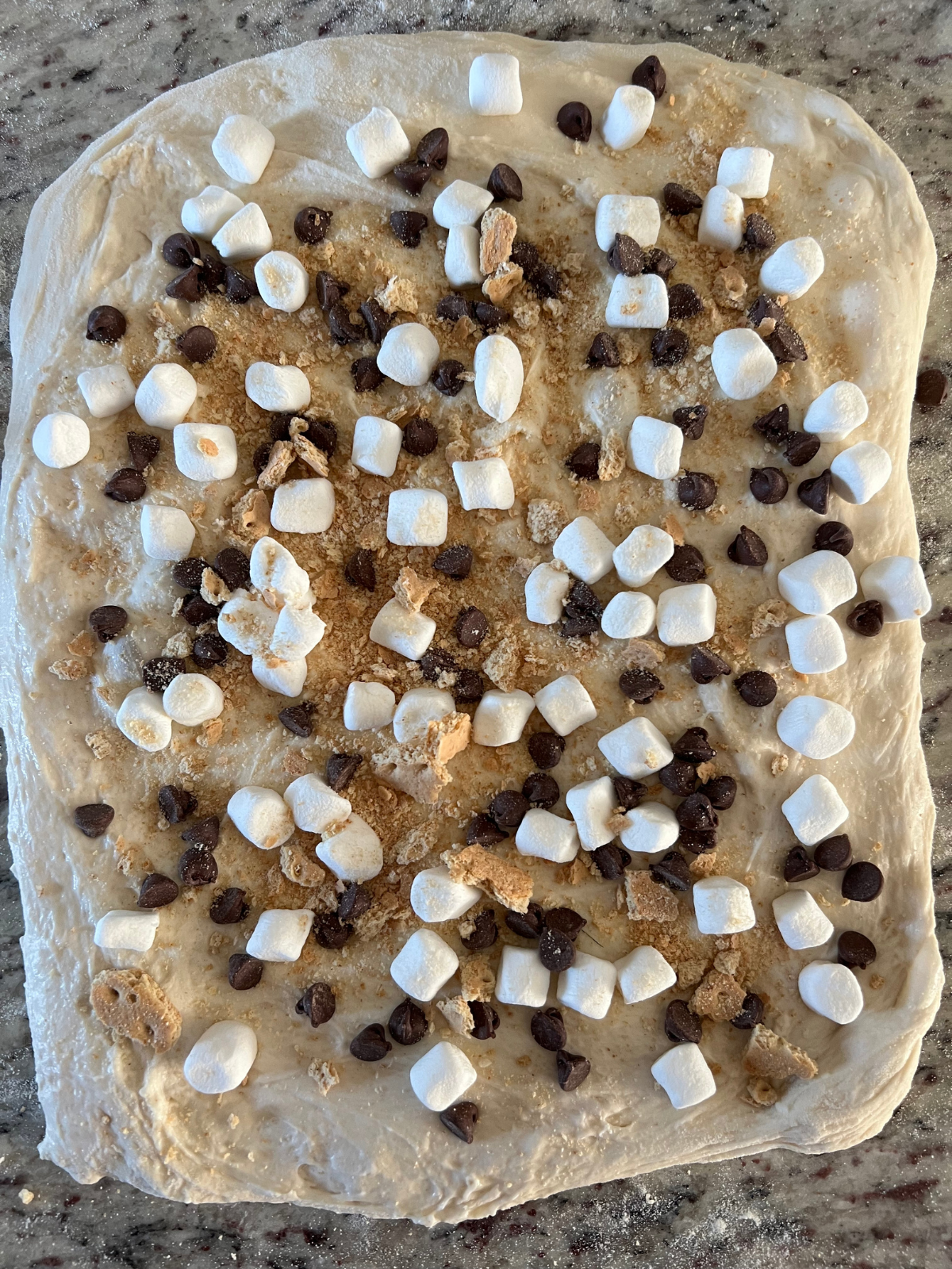 The sourdough dough has been stretched into a rectangle and the crushed graham crackers, mini marshmallows, and chocolate chips have been sprinkled over them.