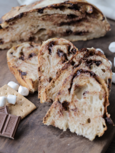 Sliced sourdough s'mores bread with chocolate chips and mini marshmallows sit with a half of the loaf in the background and marshmallows and Hershey's chocolate in the foreground.