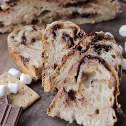 Sliced sourdough s'mores bread with chocolate chips and mini marshmallows sit with a half of the loaf in the background and marshmallows and Hershey's chocolate in the foreground.