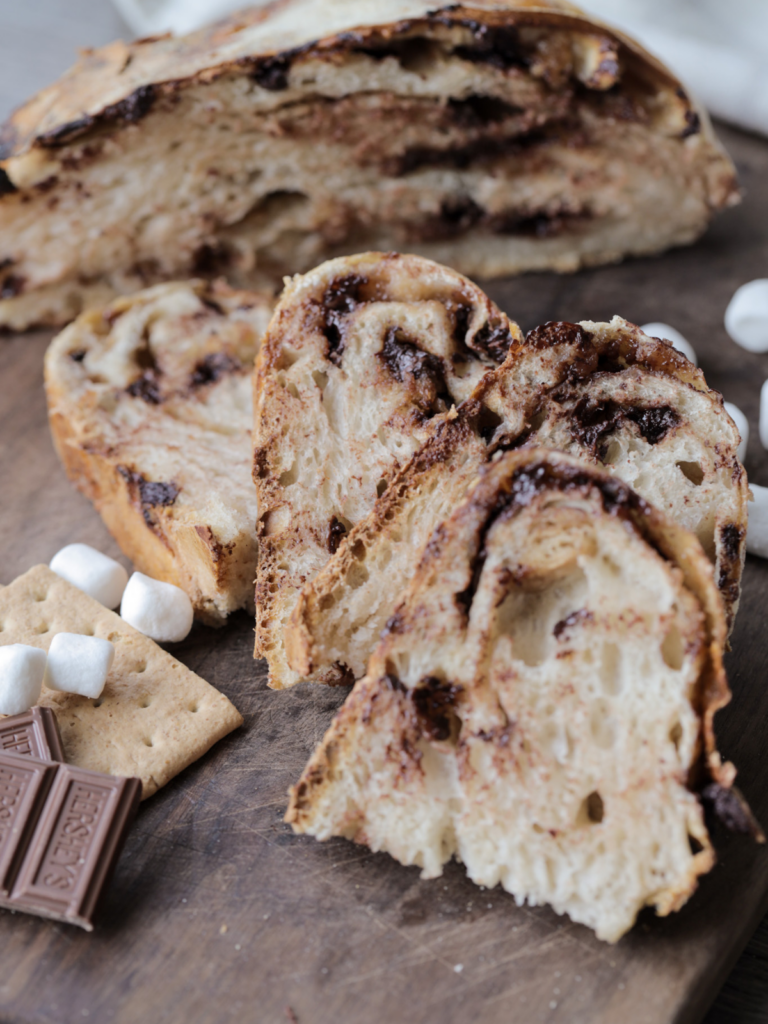 Sliced sourdough s'mores bread with chocolate chips and mini marshmallows sit with a half of the loaf in the background and marshmallows and Hershey's chocolate in the foreground.