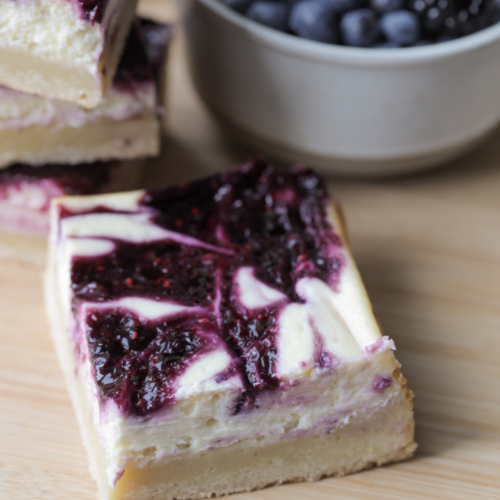 A square of berry swirl cheesecake with sourdough sugar cookie dough crust sits on a wooden cutting board with a stack of cheesecake bars and a bowl of berries in the background.