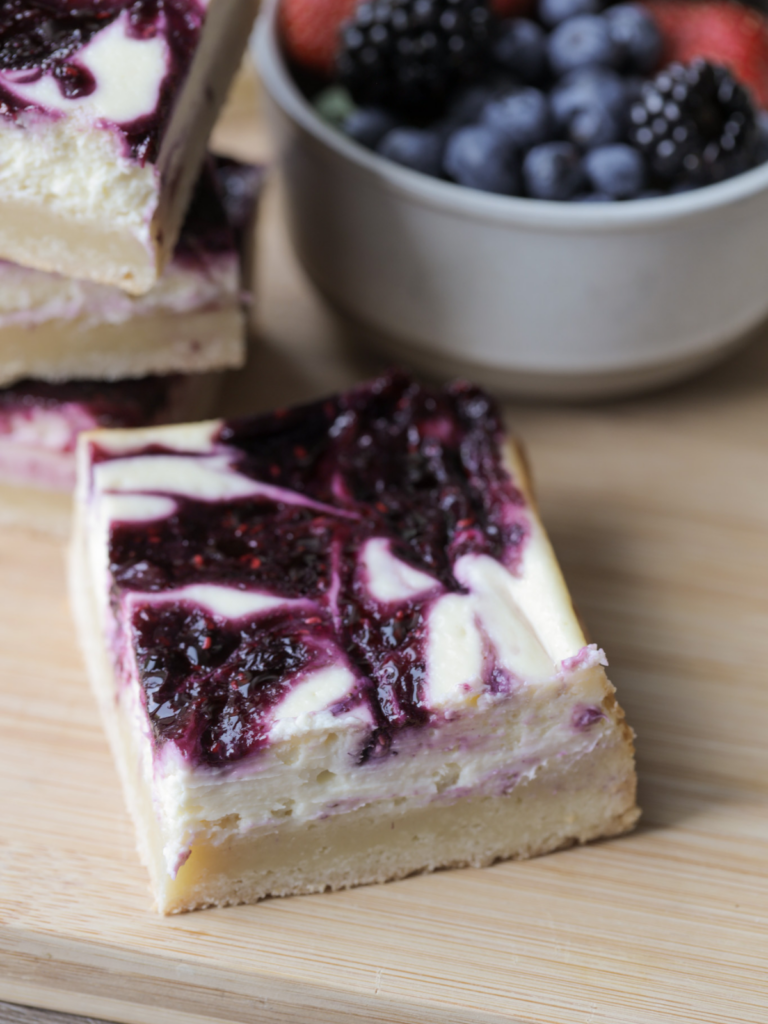 A square of berry swirl cheesecake with sourdough sugar cookie dough crust sits on a wooden cutting board with a stack of cheesecake bars and a bowl of berries in the background.