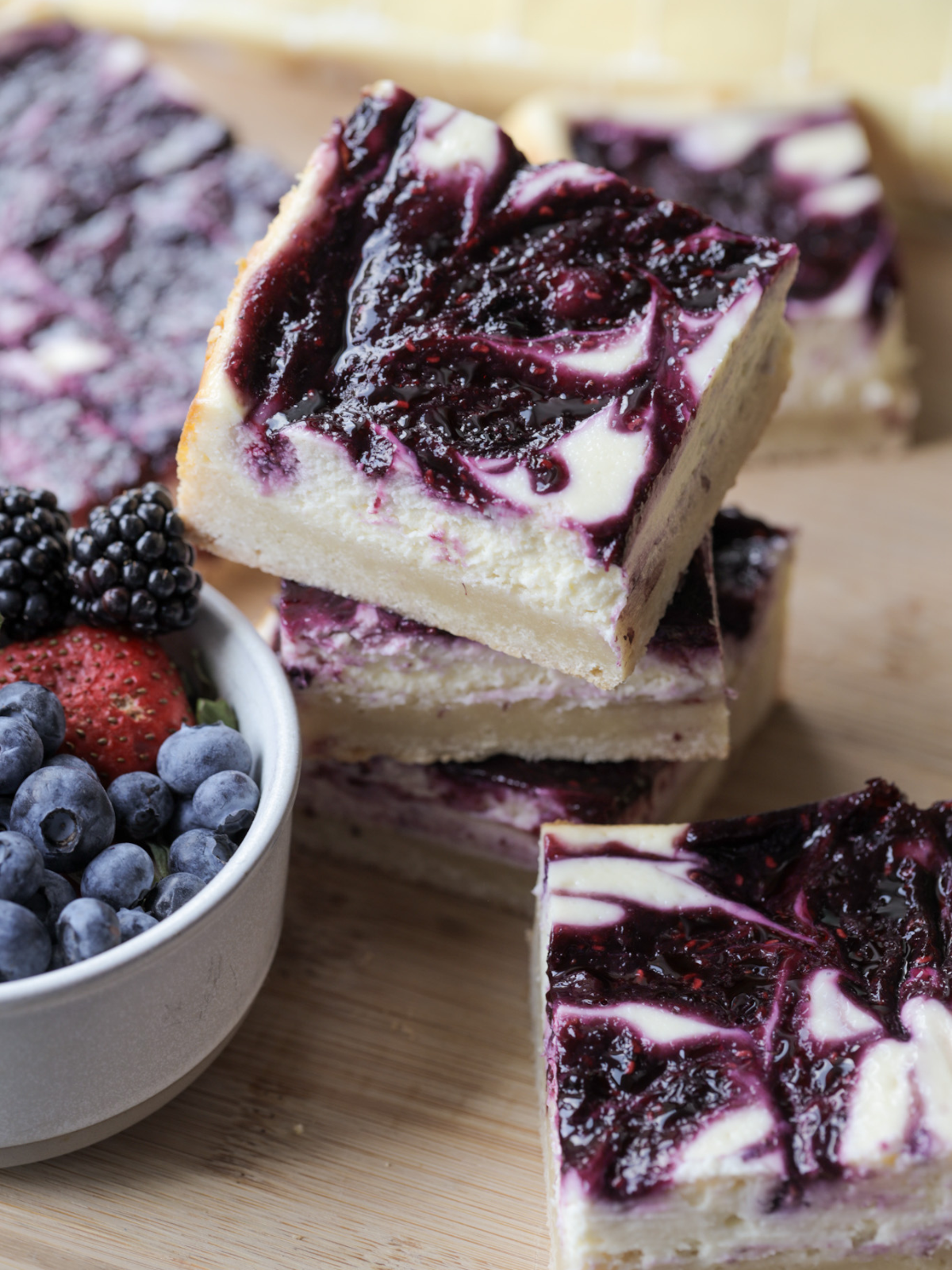 Three berry swirl cheesecake bars are shown stacked on top of each other. There are several more bars in the background along with a bowl of mixed berries.