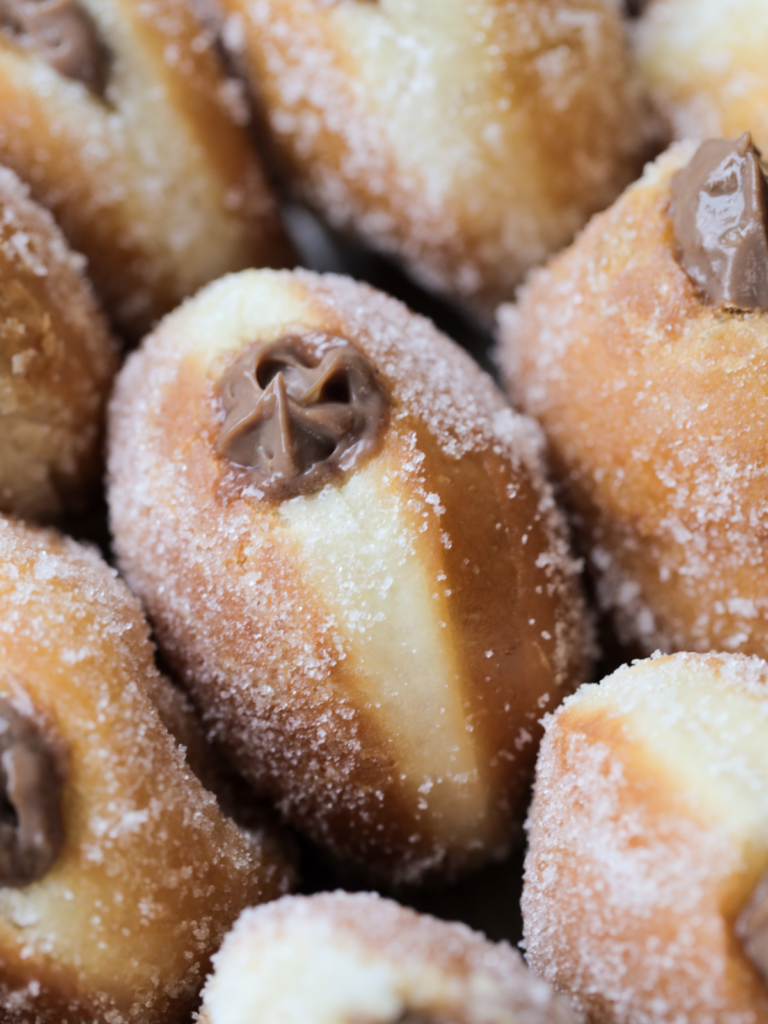 A closeup of a naturally fermented sourdough donut that is coated in granulated sugar and filled with a homemade chocolate custard. Several other donuts surround the one in focus.