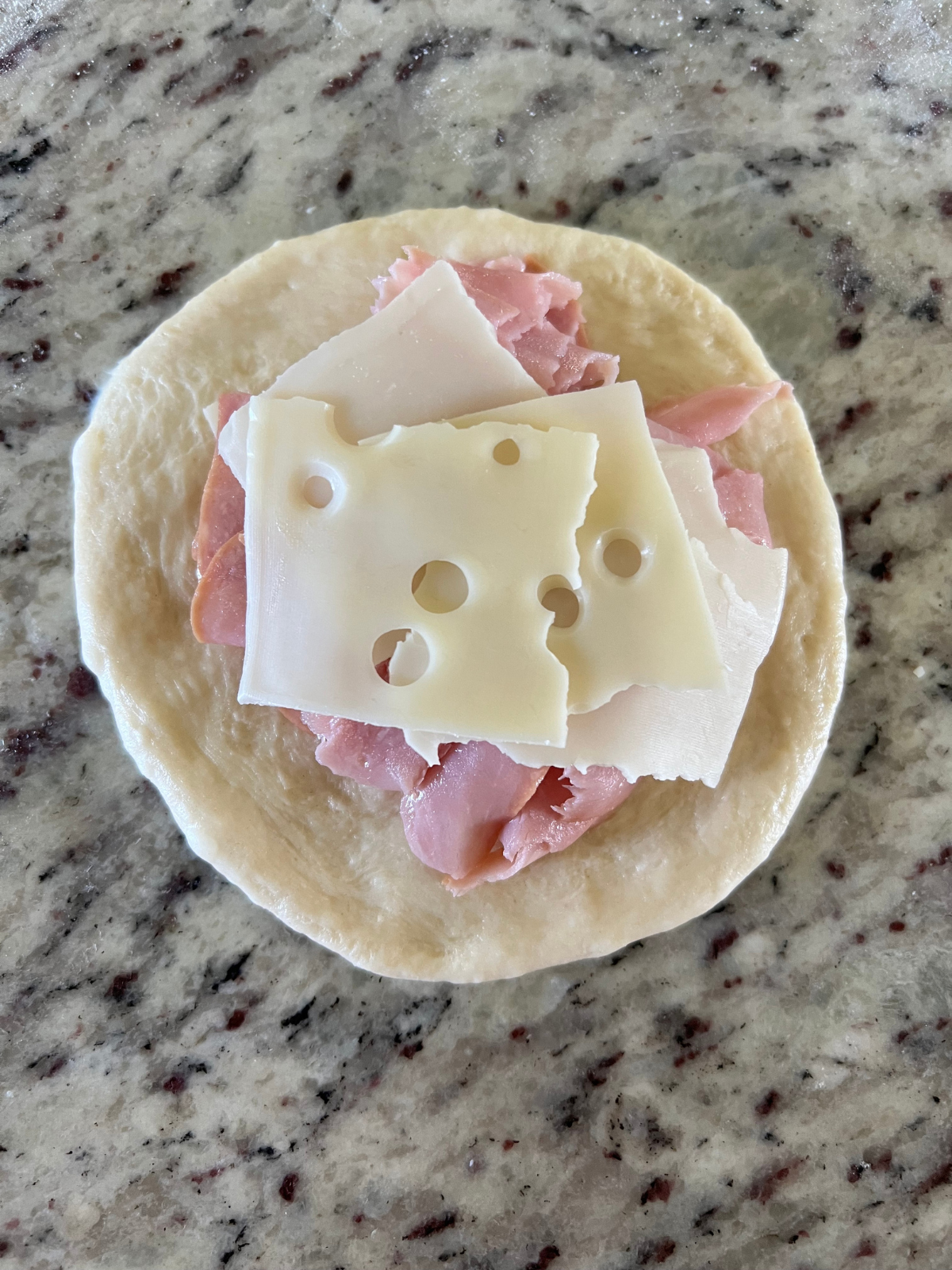 The sourdough dough ball has been flattened and topped with swiss cheese and ham.