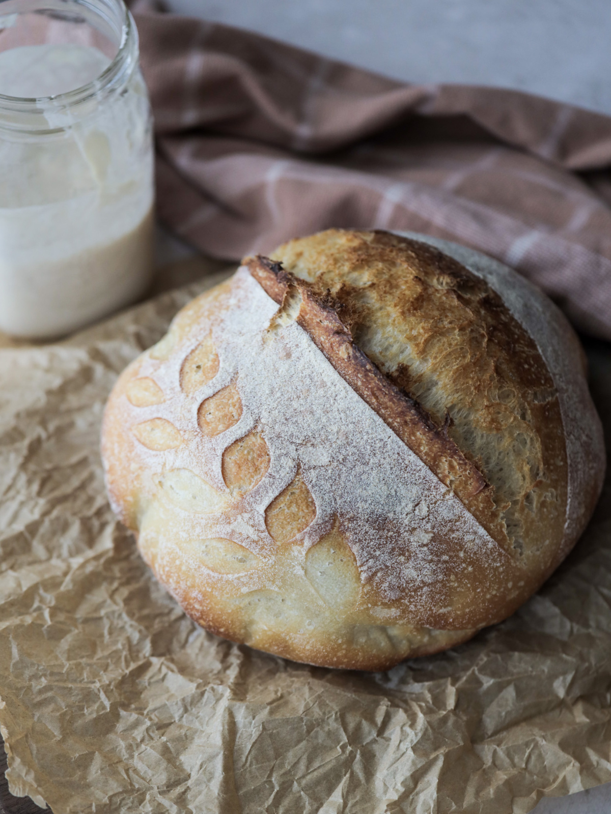 A loaf of sourdough bread is shown on parchment paper. A jar of sourdough starter sits in the background.