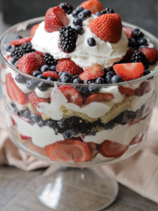 A mixed berries and cream sourdough pound cake trifle sits in a trifle dish. It's topped with whipped cream and blueberries, raspberries, strawberries, and blackberries.