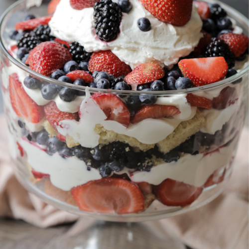 A mixed berries and cream sourdough pound cake trifle sits in a trifle dish. It's topped with whipped cream and blueberries, raspberries, strawberries, and blackberries.