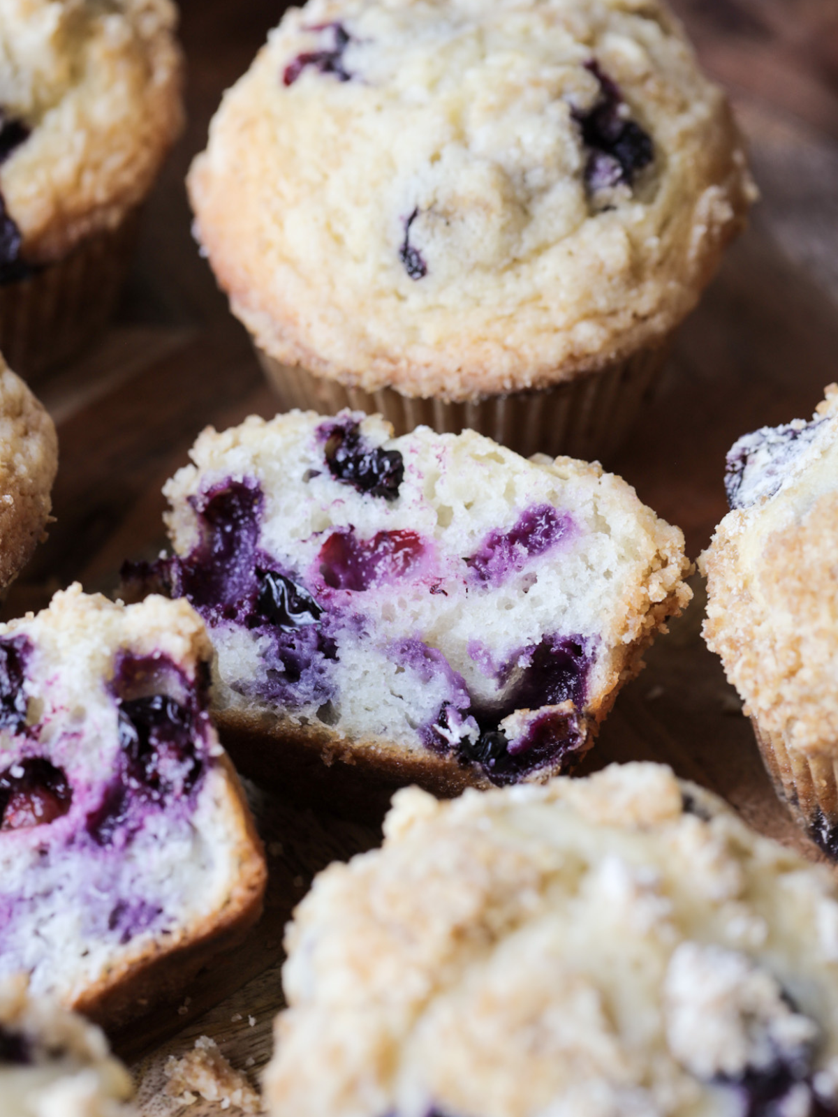 A sourdough blueberry muffin is shown in half with a burst of fresh blueberries in the center. Several other sourdough discard blueberry muffins are shown in the background and foreground.