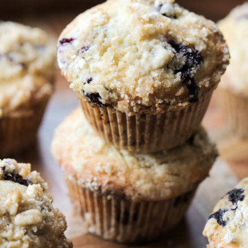 Two sourdough discard blueberry muffins are shown stacked with several other muffins in the background.