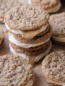 Three sourdough discard browned butter caramel apple oatmeal cream pies sit on top of each other. They have a brown sugar cream cheese frosting filling and are surrounded by several oatmeal cream pies.